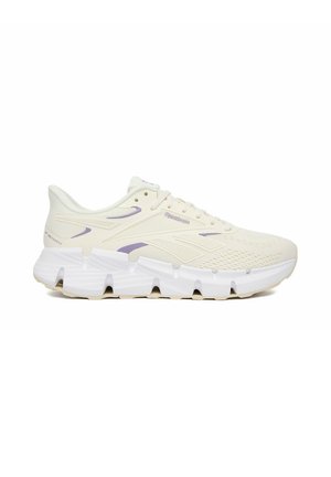 Zapatilla deportiva Reebok de color crema y púrpura claro con parte superior de malla texturizada y suela blanca gruesa segmentada, vista lateral.