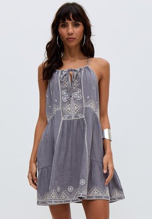 Frau trägt ein ärmelloses graues Kleid mit weißer Blumenstickerei, Schlaufenkragen und einem silbernen Armreif am linken Handgelenk.