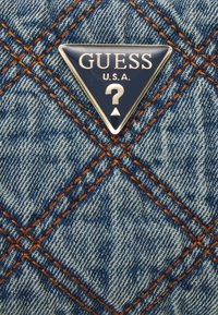 Džínová kabelka s prošívanou texturou, s oranžovým prošíváním a trojúhelníkovou modrou nášivkou s logem Guess, která má design otazníku.