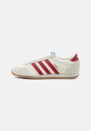 Witte Adidas sneaker met rode strepen, beige suède accenten, rubberen zool en blauw logo op de tong, zijaanzicht op een witte achtergrond.