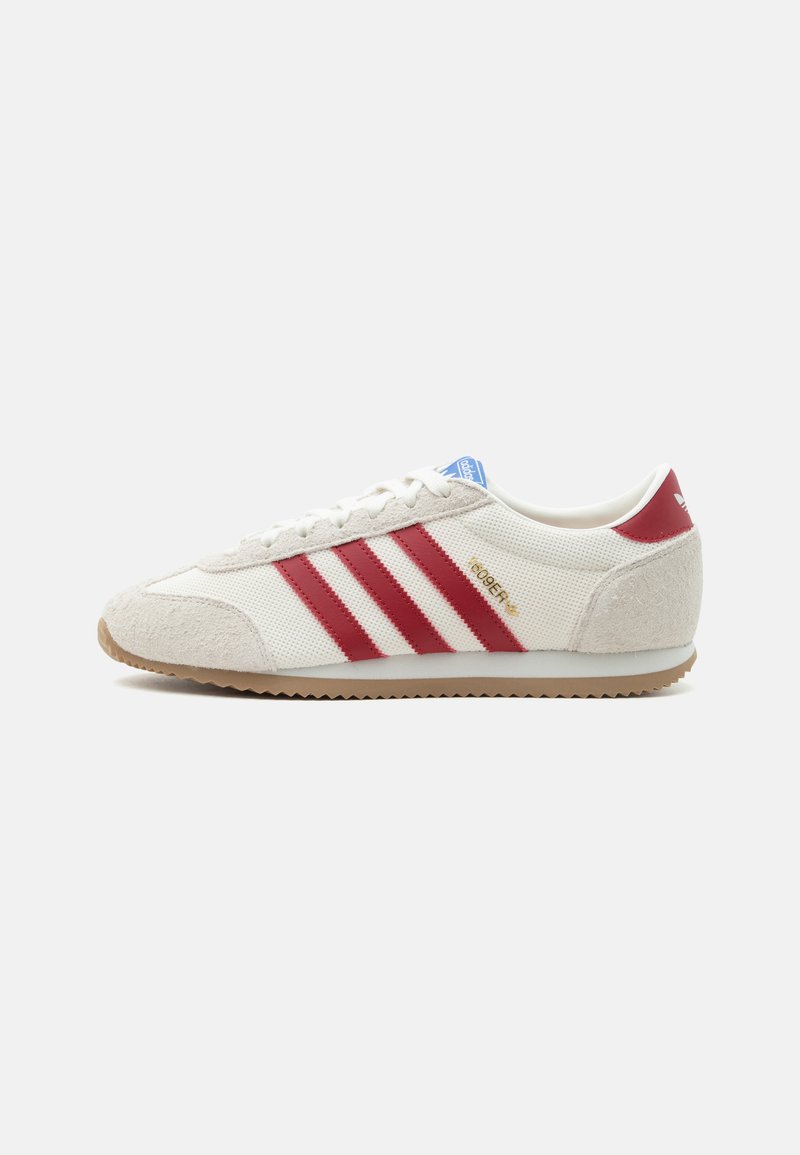 Λευκό αθλητικό παπούτσι Adidas με κόκκινες ρίγες, μπεζ σουέτ λεπτομέρειες, καουτσούκ σόλα και μπλε λογότυπο στη γλώσσα, εμφανίζεται από το πλάι σε λευκό φόντο.
