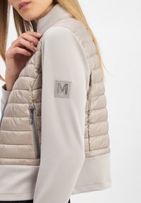 Beige gewatteerde vest met een gladde, taupe gebreide mouw en kraag. Kenmerkt zich door een zilver geborduurd 'M' embleem en een ritssluiting zijzak.