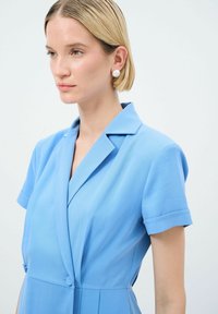 Robe portefeuille bleue avec des manches courtes, un col et deux boutons à l'avant. Texture lisse et coupe ajustée. Mannequin portant des boucles d'oreilles en perles.