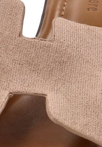 Sanda en color beige con una correa texturizada y un diseño con recortes, apoyada sobre una base de cuero marrón suave. Enfocándose en los materiales y detalles de diseño.