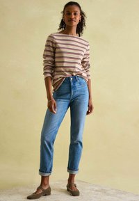 Gestreiftes langärmliges Oberteil in Beige und Kastanienbraun, kombiniert mit hellblauen High-Waist-Jeans. Das Model trägt braune Mary Jane-Schuhe und steht vor einem neutralen Hintergrund.