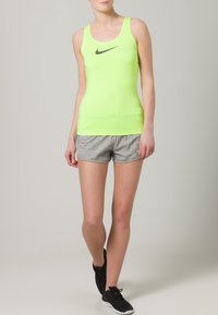 Neonowo zielony top Nike bez rękawów z czarnym logo Swoosh, zestawiony z szarymi sportowymi szortami. Materiał wygląda na gładki i dopasowany. Całość dopełniają czarne sneakersy.