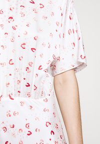 Robe blanche à manches courtes avec un imprimé floral abstrait rouge et rose dispersé et un détail élastique à la taille sur le torse d’une personne.