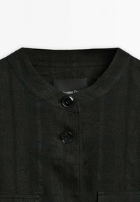Zwarte linnen blouse met een ronde halslijn, voorzien van twee zwarte knopen en een gestructureerd oppervlak. Inclusief een label aan de kraag.