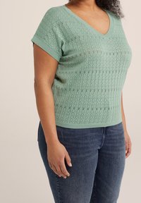 Top de punto verde claro con escote en V, mangas cortas y patrón calado texturizado. Combinado con vaqueros azul oscuro.