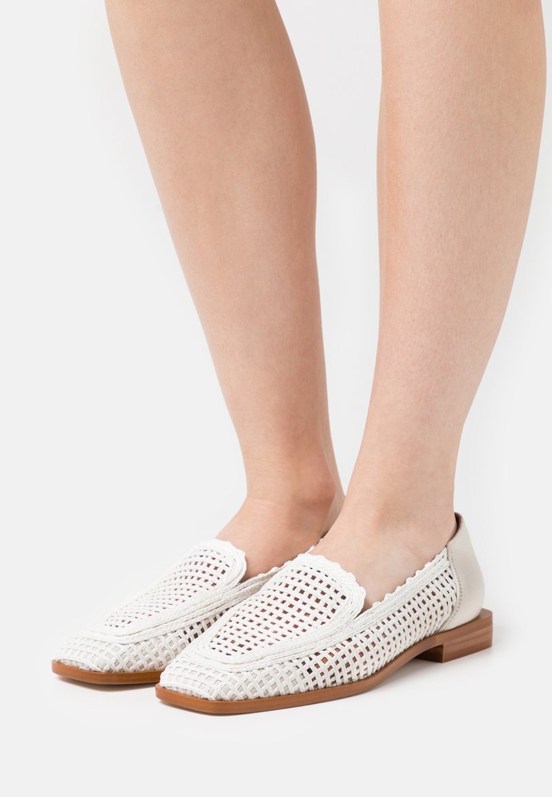 Pieds portant des mocassins blancs en cuir tressé avec des talons plats en bois sur un fond blanc.