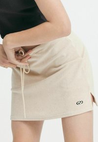 Fustă bej cu o textură moale, având slituri laterale și un șnur reglabil. Include un mic logo negru pe marginea inferioară din față.