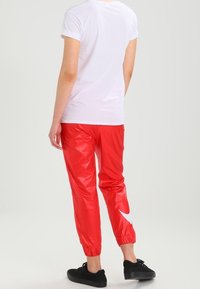 Pantalones deportivos de nailon rojos con puños elásticos y un logo blanco en el lado izquierdo, combinados con una camiseta blanca de manga corta y zapatos negros.