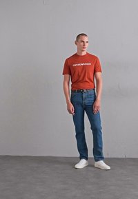 Vīrietis stāv pie pelēkas sienas, ģērbies rūsas krāsas Emporio Armani T-kreklā, zilās džinsās ar melnu jostu un baltos sporta apavos.
