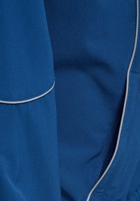 Blå jacka med ett texturerat tyg och ett subtilt ribbat mönster, grå accenter längs dragkedjorna och kanterna.