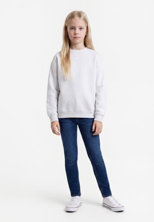 Jeune fille aux longs cheveux blonds debout portant un sweat-shirt blanc, un jean bleu et des baskets blanches sur un fond uni.