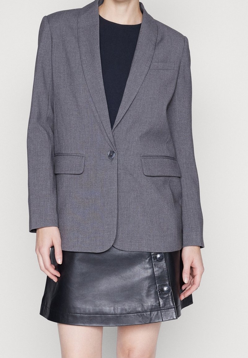 Personne portant un blazer gris par-dessus un haut noir et une mini-jupe en cuir noire avec des détails de boutons sur le côté.