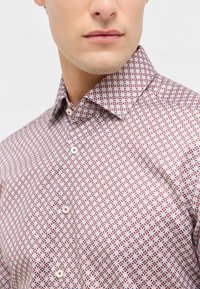 Camicia bianca con un motivo di piccoli quadrati rossi e blu. Caratterizzata da un collo a punta e una pattina frontale con bottoni bianchi.