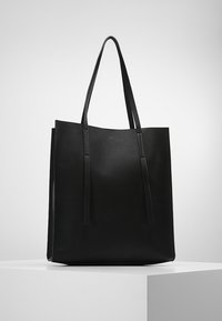 Sac en cuir noir avec deux longues poignées, en forme rectangulaire, se tenant debout sur un socle blanc contre un fond gris clair uni.