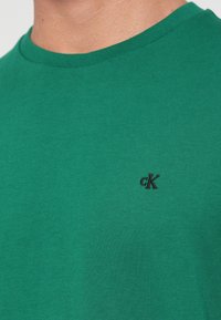 Artimas plano žmogaus su žaliu apykakle marškinėliais, ant krūtinės siuvinėtas mažas juodas "cK" logotipas.