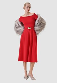 Vestito rosso scollato con corpetto aderente, gonna svasata e fibbia della cintura dorata. Abbinato a uno scialle crema peloso e tacchi beige.