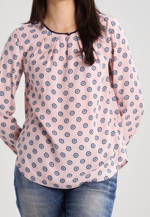 Kvinde iført en lyserød langærmet bluse med blå blomsterprint og blå jeans, står med hænderne bag ryggen.