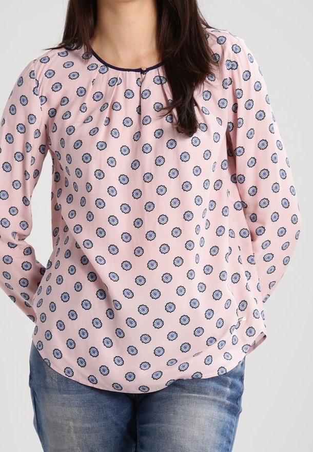 Femme portant une blouse rose pâle à manches longues avec des motifs floraux bleus et un jean bleu, debout avec les mains derrière le dos.