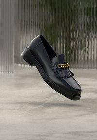 Mocassin en cuir noir avec un design à franges et un accent de chaîne en or, présentant un bout arrondi et un talon carré bas. Texture lisse.