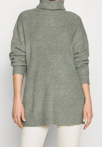 Jersey de cuello alto verde de punto con mangas largas y un corte relajado. Los puños y el dobladillo acanalados añaden textura a la prenda.