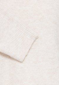 Gros plan d'une manche de pull en maille douce de couleur crème avec un détail de poignets côtelés sur un fond assorti.