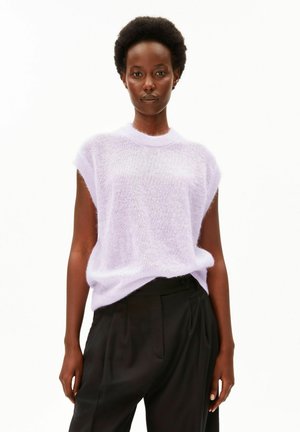 Femme aux cheveux courts et bouclés portant un haut en tricot sans manches violet clair et un pantalon noir taille haute, debout sur un fond blanc.