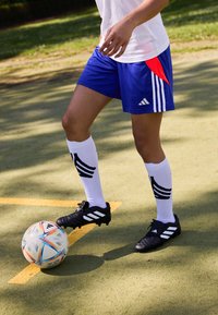Zili sporta šorti ar sarkaniem akcentiem, baltas zeķes ar melnām svītrām un melni futbolā čības ar baltām svītrām, novietoti blakus košam futbolam.