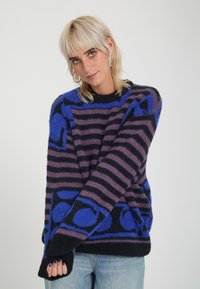 Personne aux cheveux blonds courts portant un pull rayé noir, violet et bleu avec des motifs géométriques et un jean bleu clair.