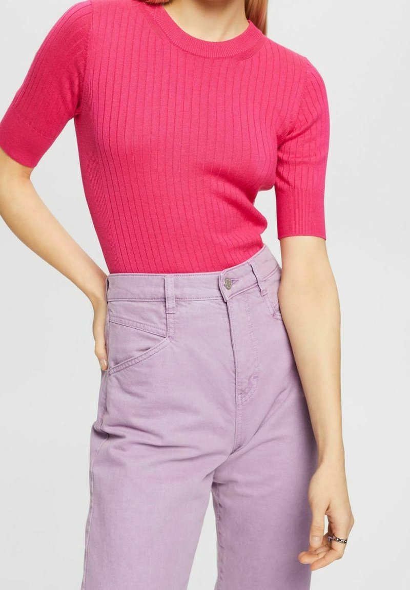 Femme portant un haut rose côtelé à manches courtes, rentré dans un jean taille haute violet clair, debout avec une main sur la hanche.