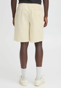 Shorts beige clair avec une taille élastique, longueur au genou, en coton. Portés avec des baskets blanches et des chaussettes grises à côtes. Design simple.