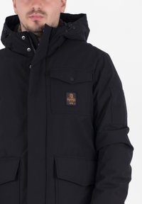 Chaqueta negra con capucha hecha de una tela suave, con un bolsillo en el pecho con tapa de botón y logo, y dos grandes bolsillos frontales.