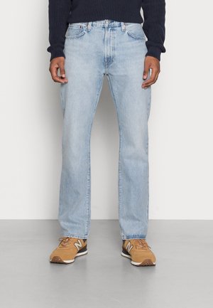 Man wearing light blue straight-leg jeans, navy sweater, and tan sneakers standing on gray floor against a plain white wall.