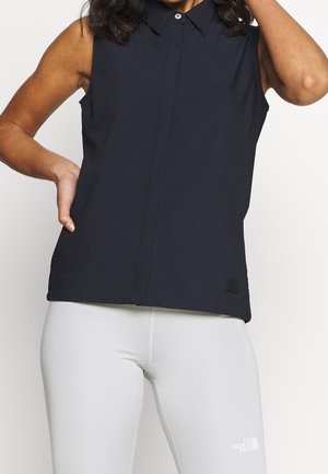 Vrouw draagt een zwarte mouwloze blouse met kraag en witte North Face legging, poseert met één hand op de heup en de andere raakt het haar aan.