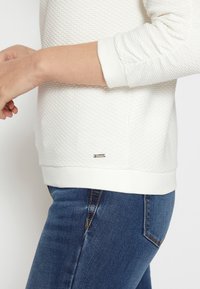 Felpa bianca con texture, maniche lunghe, polsini a costine e un piccolo tag metallico nella parte inferiore, abbinata a jeans blu scuro.