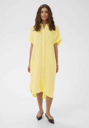 Ženska s kodrastimi temnimi lasmi, oblečena v ohlapno rumeno obleko z gumbi do kolen in črne konične čevlje, stoji pred preprostim ozadjem.