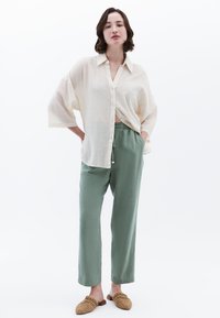 Camicia leggera beige con bottoni, dalla vestibilità ampia e maniche larghe, abbinata a pantaloni verdi rilassati e scarpe slip-on in pelle intrecciata marroni.