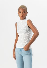 White sleeveless top with a textured, wavy pattern, high neckline, and fitted design, paired with light blue jeans.