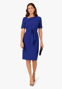 Królewsko niebieska sukienka z krótkimi rękawami, kopertowym fasonem, wiązaniem w talii i długością sięgającą kolan. Dobraną do niej małą kopertówką i cielistymi szpilkami.