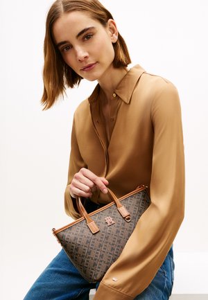 Donna con capelli castano medio alla lunghezza delle spalle, che indossa una camicetta beige e jeans blu, tenendo una borsa marrone con motivo e manici in pelle.