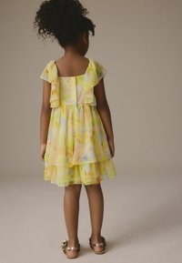Robe pour filles en tissu jaune transparent avec des motifs floraux, design en couches, manches volantes, boutons dans le dos et sandales dorées.