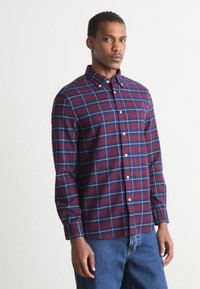 Camisa de cuadros roja y azul marino con cuello abotonado, mangas largas y botones blancos; textura suave con un corte relajado.