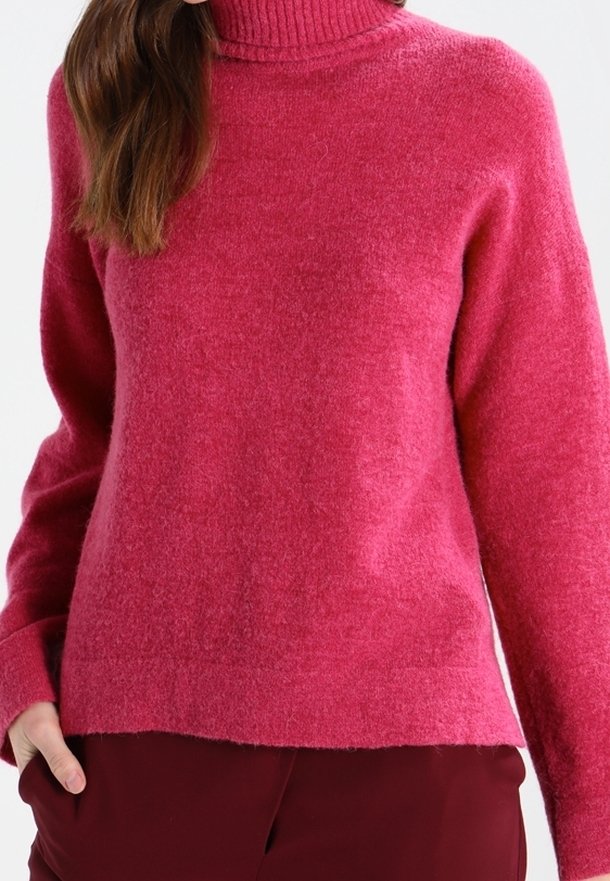Femme portant un pull rose pelucheux à manches longues et col haut, associé à un pantalon rouge foncé, debout avec une main dans la poche.