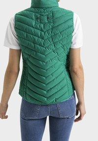 Veste matelassée verte avec un col montant, présentant un design matelassé en chevrons et des détails de boutons-pression à l'arrière. Associée à un jean bleu.