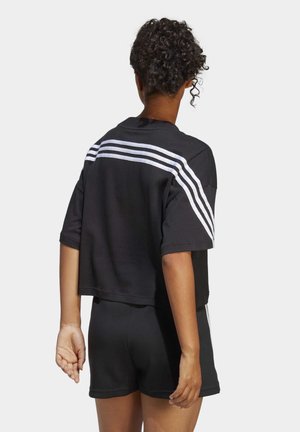 Person mit kurzen lockigen Haaren, von hinten gesehen, trägt schwarze Sportshorts und ein schwarzes Shirt mit drei weißen Streifen über den Schultern und Ärmeln.