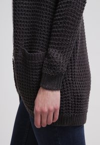 Vue latérale d'une personne portant un cardigan en maille grise foncée texturée avec une poche frontale et un jean bleu foncé.