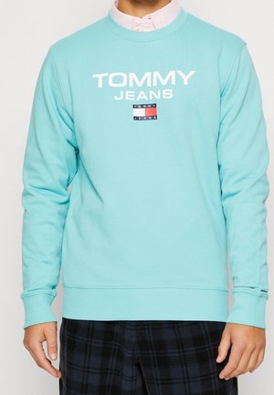 Personne portant un sweat-shirt Tommy Jeans bleu clair par-dessus une chemise col rose clair et un pantalon à carreaux foncé, debout avec les bras détendus.